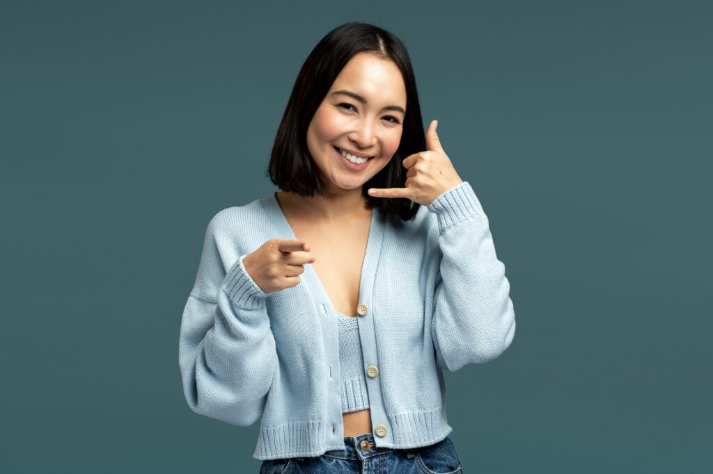 Woman standing with telephone hand gesture and smiling to camera, offering to contact by phone